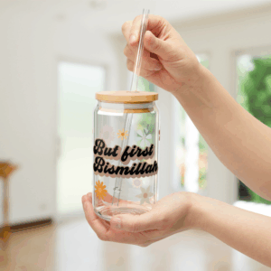 Close-up of hands holding a clear glass tumbler with a bamboo lid and reusable straw. The tumbler is decorated with colorful flower illustrations and black cursive text that reads “But first Bismillah.” The background shows a bright, softly blurred indoor setting with natural light and greenery outside.