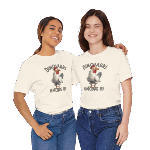 Two smiling women standing close together, each wearing an off-white T-shirt with an illustration of a cartoon chicken and the text “DINOSAURS AMONG US.” Both have one arm around each other and are dressed casually in jeans against a plain black background.