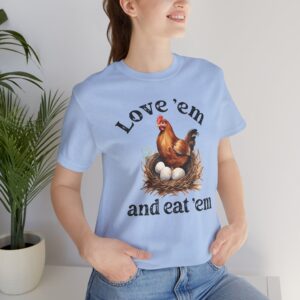 Woman wearing a light blue t-shirt featuring an illustration of a brown hen sitting in a nest with three white eggs. The text above and below the image reads “Love ’em and eat ’em” in a bold, playful black font. She is smiling and standing casually with her hands in her pockets, and there is a green potted plant in the background.