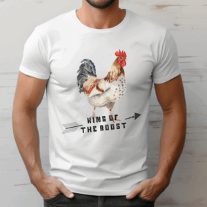 Man wearing a white t-shirt featuring a watercolor-style illustration of a rooster with red, brown, and black feathers. Below the rooster, the text reads “King of the Roost” in bold, rustic lettering with a black arrow graphic running through it. The man stands casually with his hands in his pockets against a light, textured background.